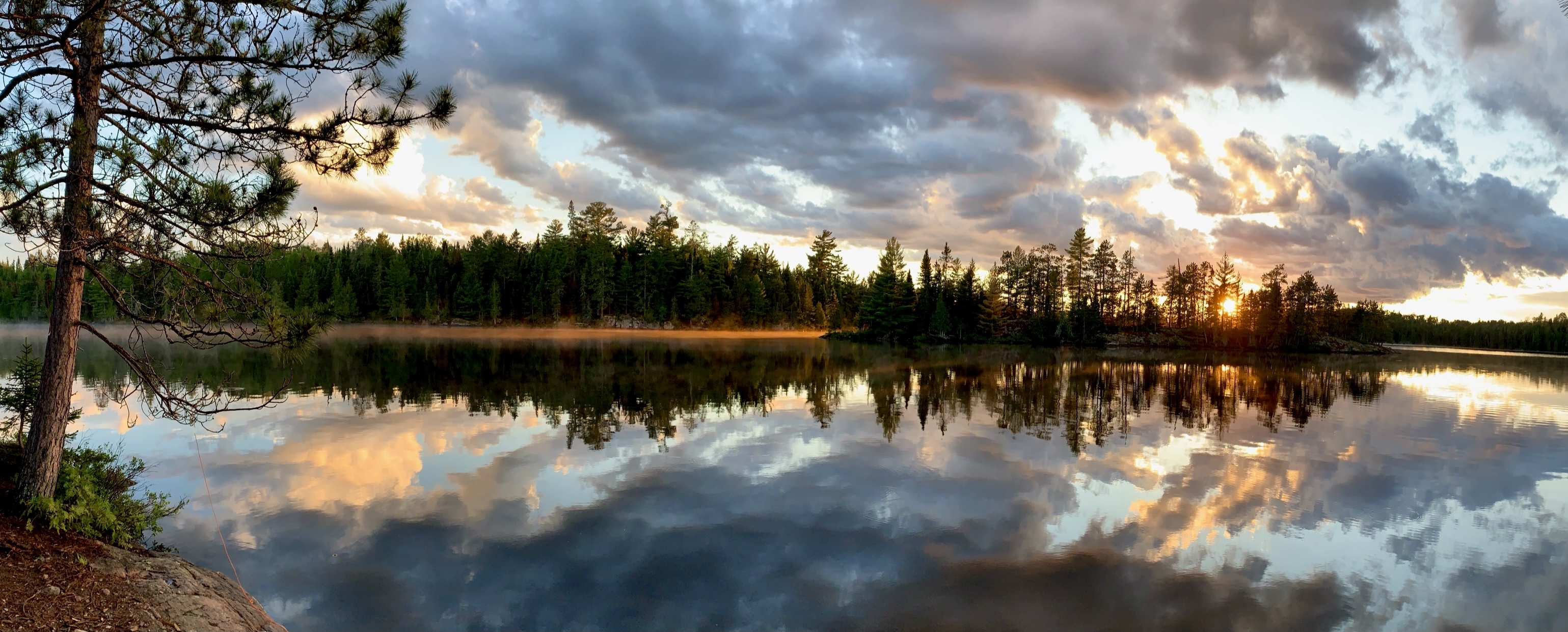Panoramic view of a calm Canadian lake at sunset with dramatic clouds and a pine forest reflected in the still water.
