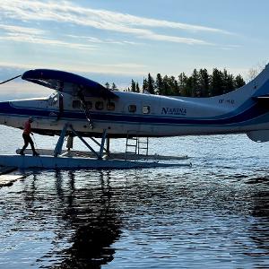 A blue and white Nakina Air Service Otter floatplane docking at a wooden pier at their airbase with a person stepping onto the float.
