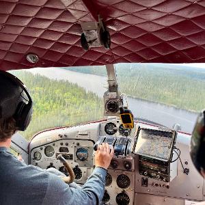 Interior view of a bush plane cockpit showing the pilot, instrument panel, and a view of the boreal forest and river below.