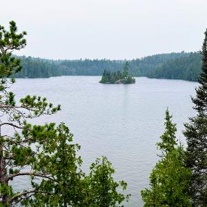A wide lake surrounded by evergreen trees with a small forested island in the center under a cloudy sky.