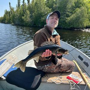 A fisherman in a small boat holds up a large walleye caught on a remote Canadian lake surrounded by dense forest.