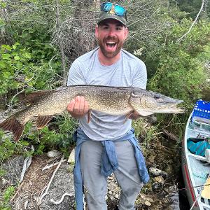 A man holding a large Northern Pike fish horizontally, smiling broadly, standing on a rocky shore next to a boat.