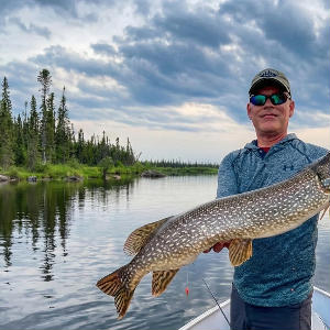 Angler holding a large Northern Pike in a boat on a remote Canadian lake under a cloudy sky.