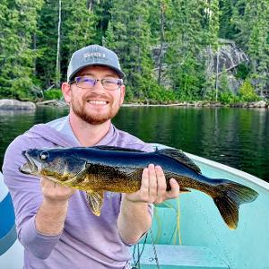 A man on a boat smiles while holding a large walleye. A dense green forest and calm lake are in the background.