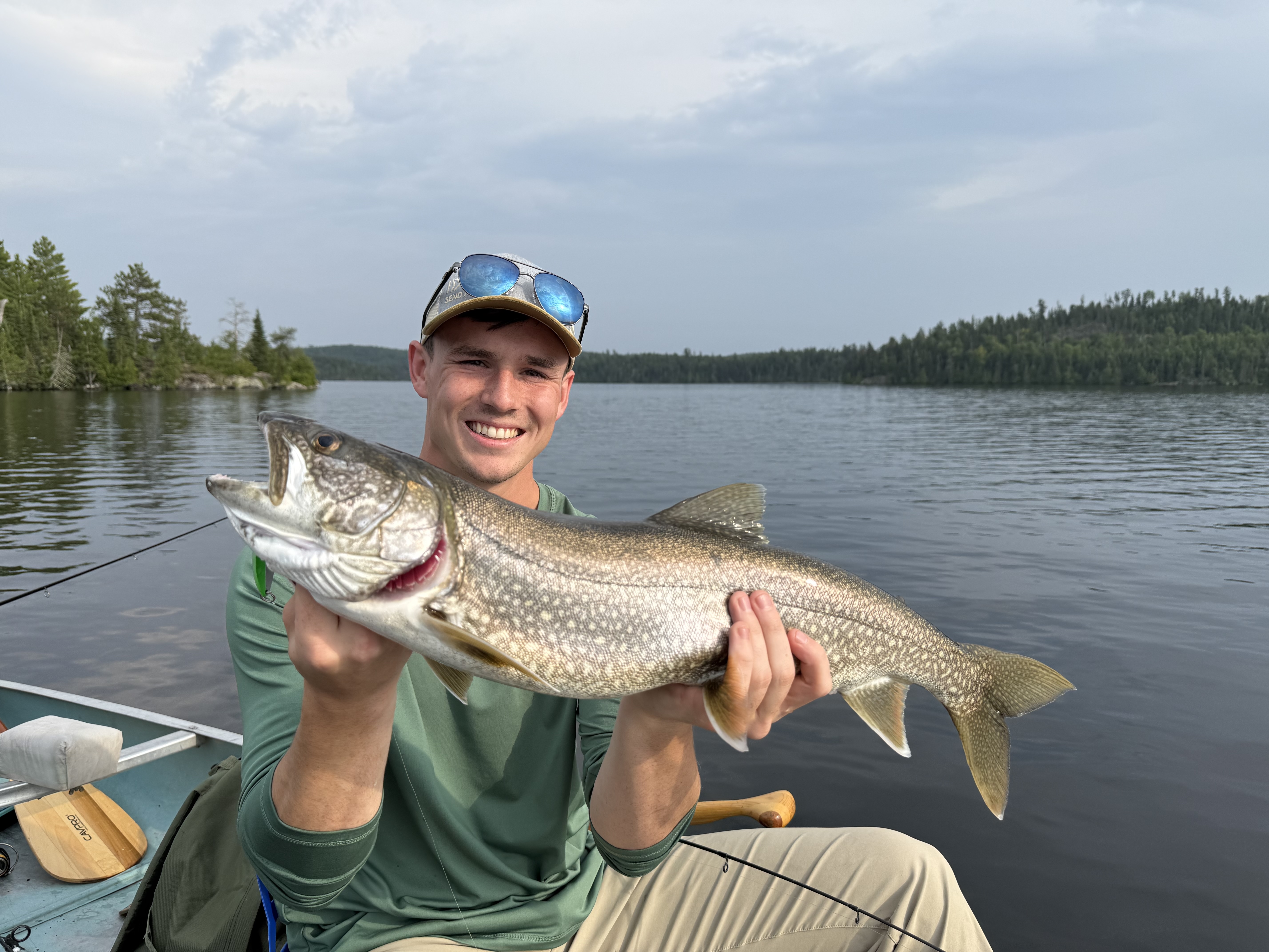 A smiling young man in a boat holding a large, spotted Lake Trout horizontally against a backdrop of a calm lake and pine forest.