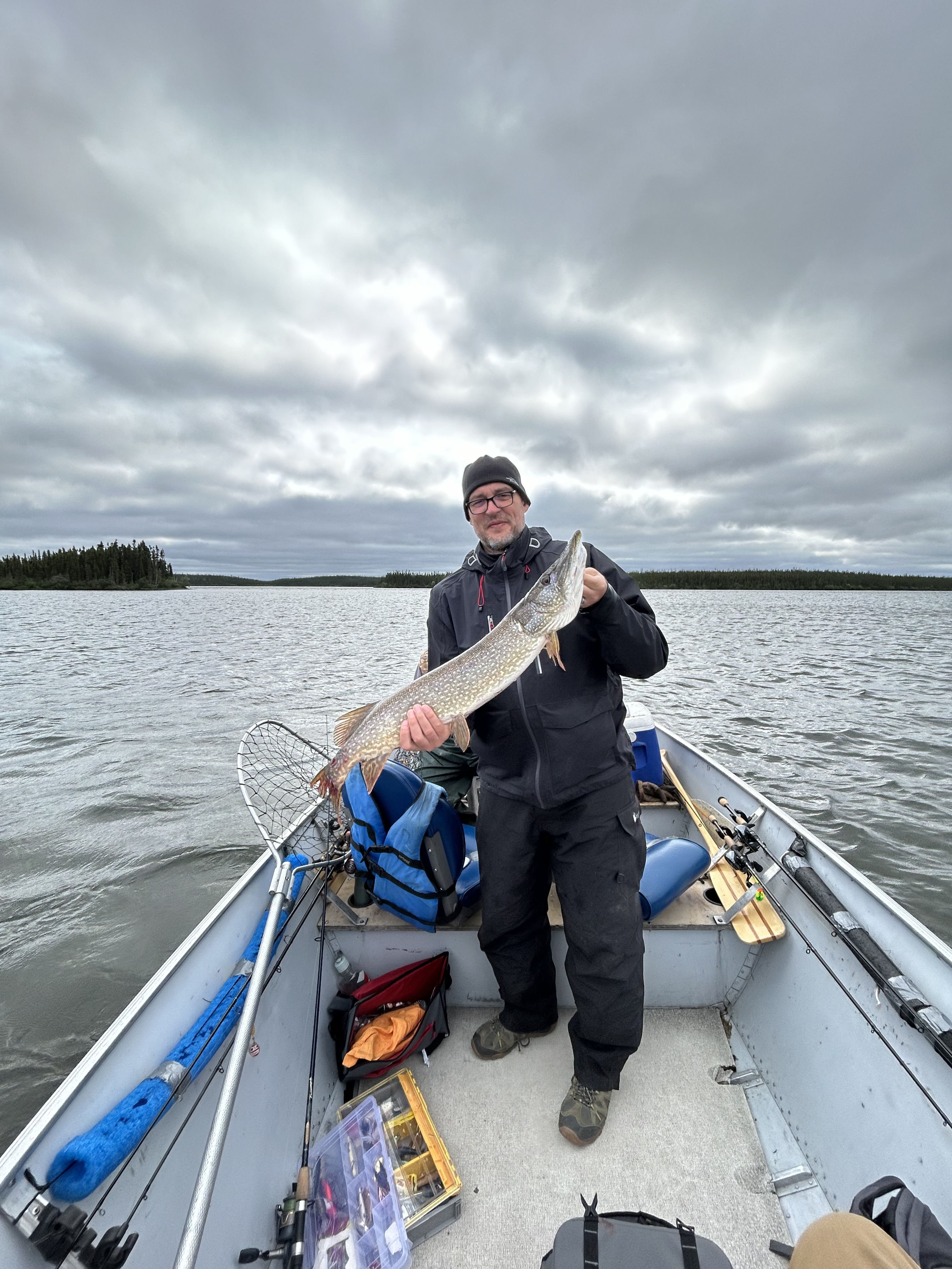 Angler holding a large Northern Pike in a boat on a remote Canadian lake under a cloudy sky.